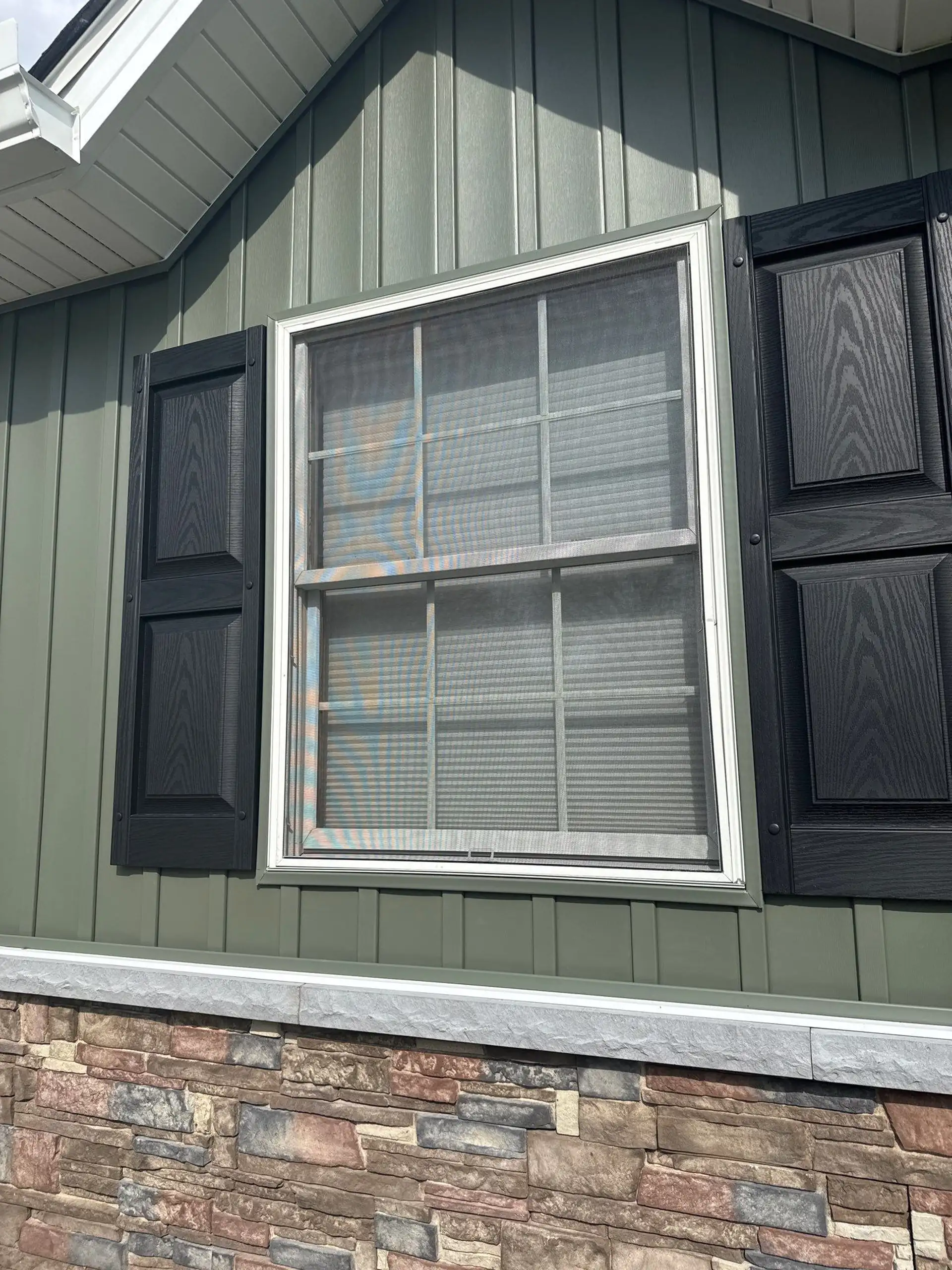 Green board-and-batten exterior siding with white-trim window, black decorative shutters and stone veneer foundation
